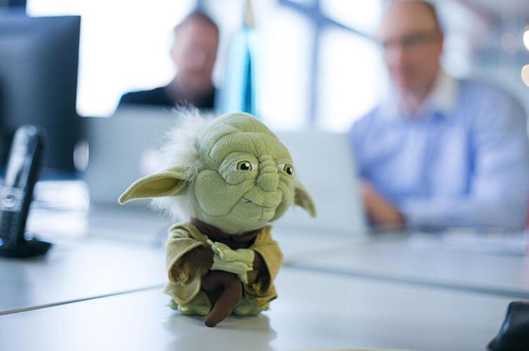 A photo of a Yoda plushie on a desk with two people in the background.